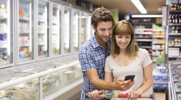 Un homme et une femme se tiennent près l'un de l'autre dans un supermarché, tandis que l'homme regarde un smartphone et la femme jette un œil par-dessus son épaule. Derrière eux, on peut voir des congélateurs et des étagères avec des produits.