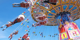 Des personnes sont assises dans des chaises berçantes d'un manège qui tourne haut dans les airs, avec en arrière-plan des magasins colorés et un ciel bleu lumineux.