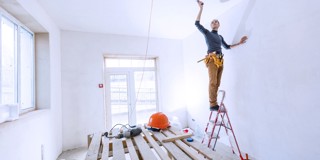 Une personne se tient sur une échelle en train de peindre un plafond blanc dans une pièce vide avec un sol en planches de bois, un casque de chantier et un chevalet visibles en arrière-plan.