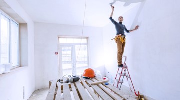 Une personne se tient sur une échelle en train de peindre un plafond blanc dans une pièce vide avec un sol en planches de bois, un casque de chantier et un chevalet visibles en arrière-plan.