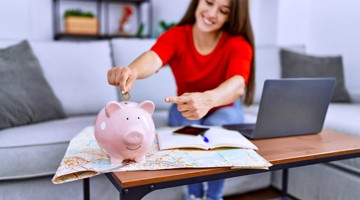 Une personne mettant des pièces dans une tirelire rose sur une table en bois avec un ordinateur portable et un carnet.