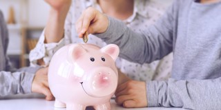 Un cochon tirelire rose avec un visage souriant, tandis qu'une main y place une pièce de monnaie, en arrière-plan, quelques personnes assises à une table.